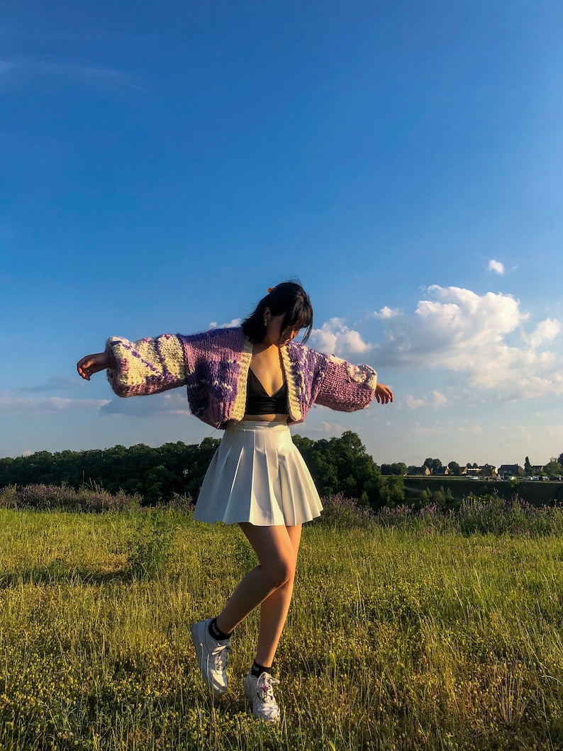 Peut inclure: Une femme portant une jupe pliss&eacute;e blanche, un soutien-gorge noir et un cardigan en tricot violet et blanc &agrave; motif floral. Elle se tient dans un champ d'herbe verte avec un ciel bleu en arri&egrave;re-plan.