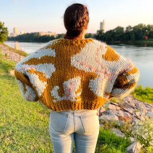 Peut inclure: Une personne portant un pull en tricot &eacute;pais avec un motif abstrait brun et blanc. Le pull a une coupe ample et est court &agrave; la taille. La personne porte un jean bleu et se tient devant une &eacute;tendue d'eau.