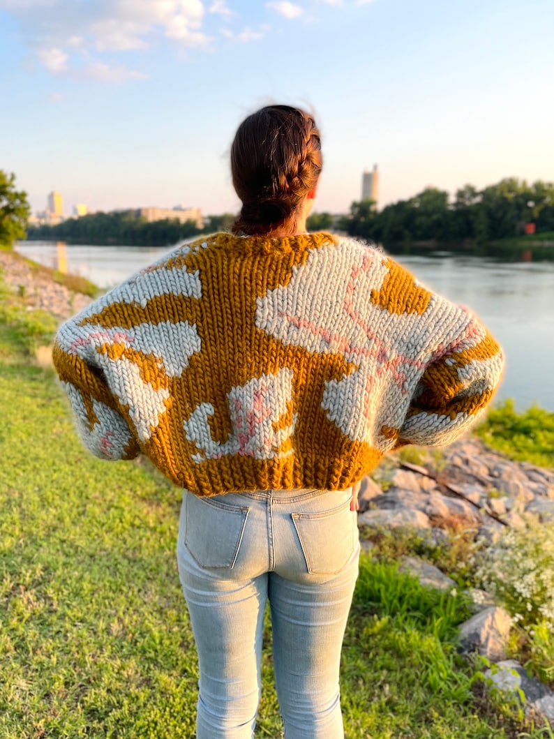 Peut inclure: Une personne portant un pull en tricot &eacute;pais avec un motif abstrait brun et blanc. Le pull a une coupe ample et est court &agrave; la taille. La personne porte un jean bleu et se tient devant une &eacute;tendue d'eau.