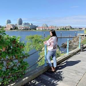 Peut inclure: Une femme portant un pull &agrave; motifs rose et blanc et un jean bleu se tient sur une passerelle en bois surplombant une &eacute;tendue d'eau. La passerelle est bord&eacute;e d'une cl&ocirc;ture en cha&icirc;ne et de rampes bleues. La femme regarde l'eau. &Agrave; l'arri&egrave;re-plan, on aper&ccedil;oit des b&acirc;timents et un ciel bleu.