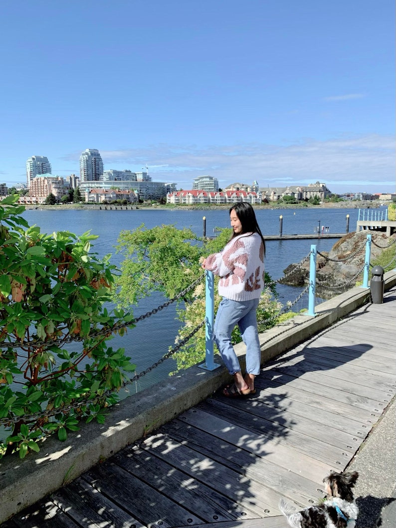Peut inclure: Une femme portant un pull &agrave; motifs rose et blanc et un jean bleu se tient sur une passerelle en bois surplombant une &eacute;tendue d'eau. La passerelle est bord&eacute;e d'une cl&ocirc;ture en cha&icirc;ne et de rampes bleues. La femme regarde l'eau. &Agrave; l'arri&egrave;re-plan, on aper&ccedil;oit des b&acirc;timents et un ciel bleu.