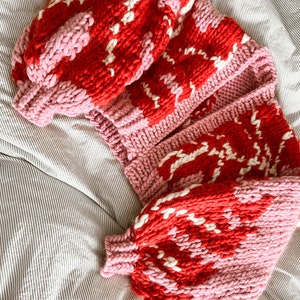 Peut inclure: Un cardigan en tricot rose et rouge avec un motif blanc. Le cardigan est pli&eacute; et pos&eacute; sur une couverture de lit ray&eacute;e.