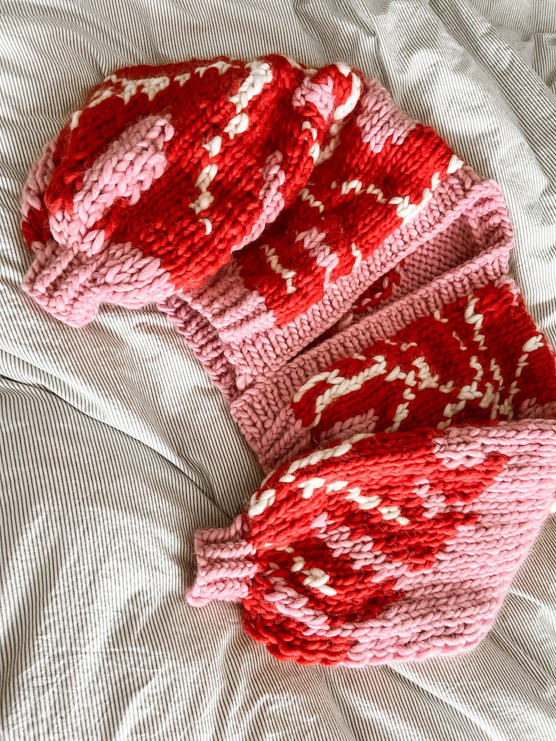 Peut inclure: Un cardigan en tricot rose et rouge avec un motif blanc. Le cardigan est pli&eacute; et pos&eacute; sur une couverture de lit ray&eacute;e.