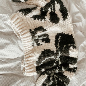 Peut inclure: Un pull en tricot cr&egrave;me avec un motif abstrait noir et blanc. Le pull est ample et a une texture tricot&eacute;e &eacute;paisse.