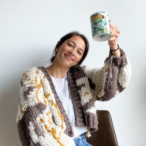 Peut inclure: Une femme portant un cardigan ray&eacute; marron, blanc et jaune avec un t-shirt blanc et un short en jean bleu tient une tasse en c&eacute;ramique blanche avec un motif de chat de dessin anim&eacute;. La tasse porte le texte "The Cat in the Hat".