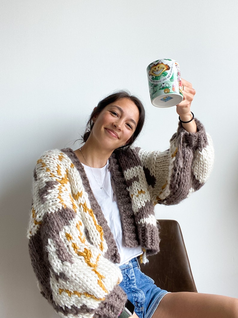 Peut inclure: Une femme portant un cardigan ray&eacute; marron, blanc et jaune avec un t-shirt blanc et un short en jean bleu tient une tasse en c&eacute;ramique blanche avec un motif de chat de dessin anim&eacute;. La tasse porte le texte "The Cat in the Hat".