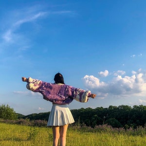 Peut inclure: Une personne portant un pull en tricot violet et blanc avec une jupe pliss&eacute;e blanche, debout dans un champ d'herbe verte et un ciel bleu.