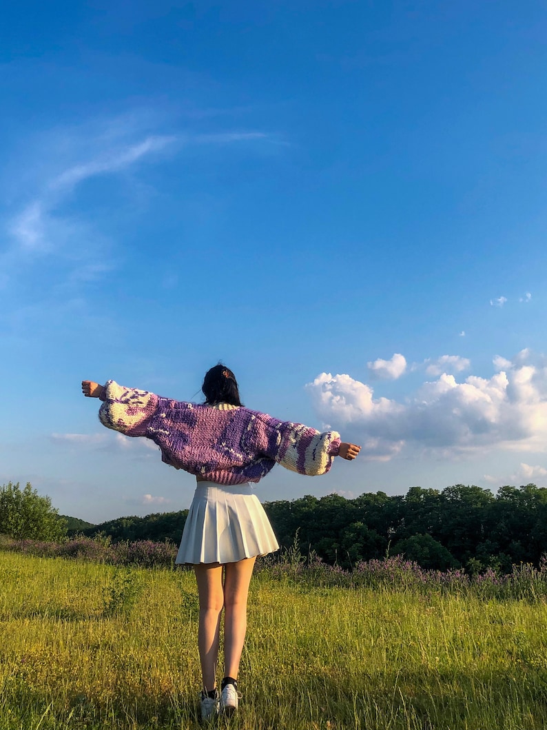 Peut inclure: Une personne portant un pull en tricot violet et blanc avec une jupe pliss&eacute;e blanche, debout dans un champ d'herbe verte et un ciel bleu.