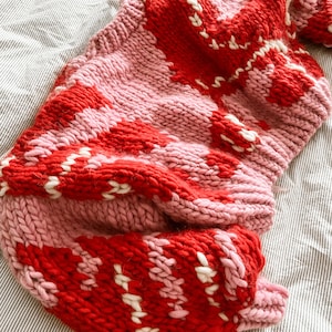 Peut inclure: Une couverture en tricot rose et rouge avec un motif g&eacute;om&eacute;trique. La couverture est faite de fil &eacute;pais et a une texture douce et confortable.