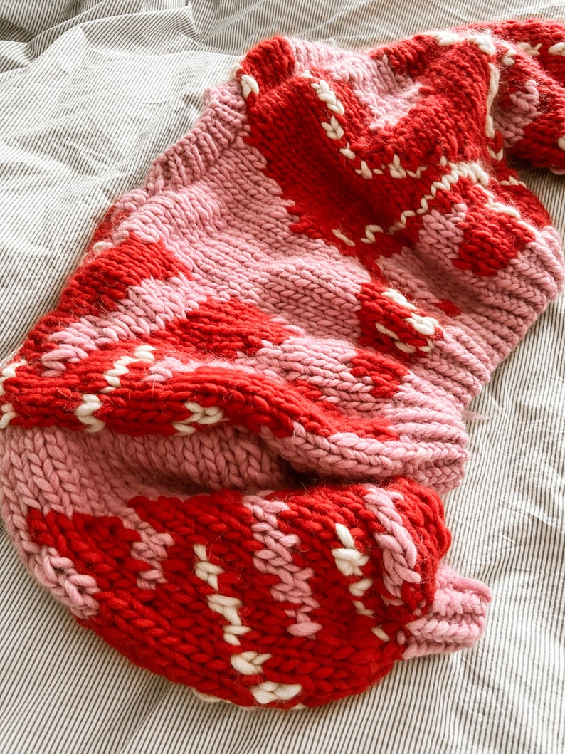 Peut inclure: Une couverture en tricot rose et rouge avec un motif g&eacute;om&eacute;trique. La couverture est faite de fil &eacute;pais et a une texture douce et confortable.