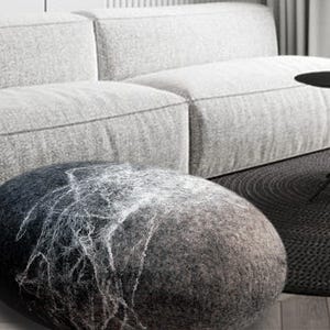 Könnte beinhalten: Ein großer, runder, grauer und weißer Filz-Pouf. Der Pouf steht auf einem schwarz-weißen gemusterten Teppich vor einem grauen Sofa.