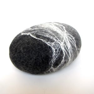 Könnte beinhalten: Ein dunkelgraues, oval geformtes Filzseifenstück mit einem weißen, netzartigen Design auf der Oberseite. Die Seife hat eine glatte, abgerundete Form und ein natürliches, handwerkliches Aussehen. Der Hintergrund ist weiß.