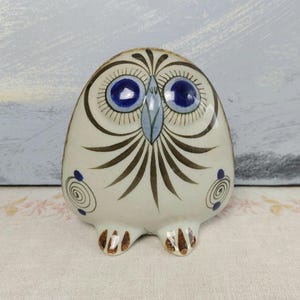 May include: A ceramic owl figurine with a white base, featuring blue eyes, brown and black feather details, and small brown feet. The owl has a whimsical design with spiral accents on its sides. The background is a light blue and white.