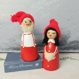 May include: Two wooden Christmas figurines. One figure is dressed in a red and white outfit with a red hat and a white beard. The other figure is dressed in a red dress and a red hat. The figurines are on a blue book that says "Merry Christmas".