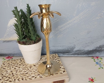 Puede incluir: Un candelero de metal dorado con forma de piña y diseño de palmera. El candelero está sobre un libro con un patrón floral.