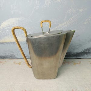 Op de afbeelding: Een theepot van geborsteld metaal met een geweven handvat en dekselhandvat. De theepot heeft een rechthoekige body en een tuit aan de zijkant. Het handvat is gemaakt van een lichtbruin geweven materiaal. De achtergrond is lichtblauw en wit.