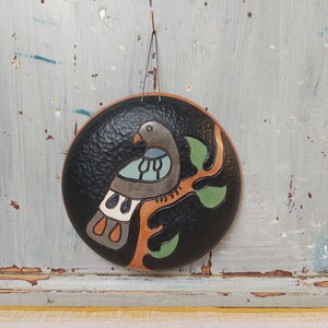 May include: A circular black wall plaque featuring a textured surface and a central illustration of a bird perched on a branch. The bird is depicted in shades of gray, light blue, and white, while the branch is orange with green leaves.