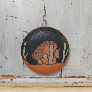 Puede incluir: Una placa de pared circular de cerámica con un diseño en relieve de un pez nadando en un mar oscuro y texturizado. El pez está representado en tonos naranja y gris, sobre un fondo negro con detalles de algas verde claro y una base de terracota.