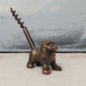 Puede incluir: Un sacacorchos con forma de perro de color bronce. El perro está de pie, con un sacacorchos que se extiende hacia arriba. El metal tiene un aspecto texturizado. El fondo es un azul claro y blanco borroso.