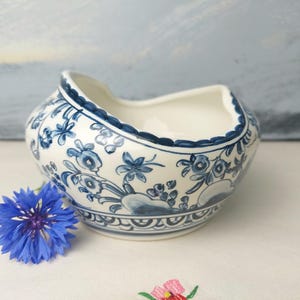May include: A white ceramic bowl with a blue floral design and a scalloped edge. The bowl has a distinctive curved opening. A blue flower sits beside the bowl, adding colour. The bowl is likely for decorative purposes, perhaps for display.