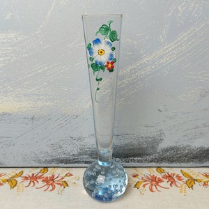 May include: A clear glass bud vase with a blue, white and red painted flower design. The vase has a round, textured base.
