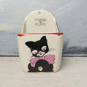 Puede incluir: Un bolsillo de pared de cerámica vintage con una ilustración de un gato negro con un lazo rosa. El bolsillo tiene un borde rojo y el texto "TREKK of KLIPP" está impreso en la parte superior. El gato tiene una expresión gruñona.