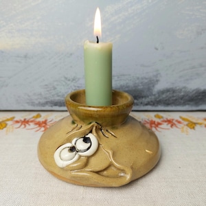 May include: A ceramic candle holder with a whimsical face and large eyes. The holder is glazed in a light brown colour and has a green candle inside.