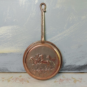 Può includere: Una padella color rame, in stile vintage, con un lungo manico e un anello all'estremità. La padella rotonda presenta un rilievo dettagliato di tre cavalli. La padella è probabilmente utilizzata per scopi decorativi.
