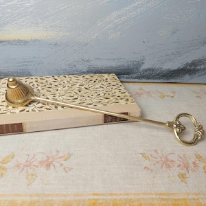 May include: A gold-toned metal candle snuffer with a decorative handle. The snuffer is resting on a book with a patterned cover.