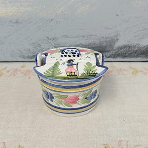 May include: A small, hand-painted ceramic container with a lid. The container is white with blue, yellow, and pink floral designs. The lid features a painted scene of a person with a staff. The container has two small handles.