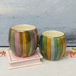 May include: Two ceramic mugs with striped patterns. The mug on the left has pink, yellow, and green stripes. The mug on the right has yellow, blue, and green stripes.