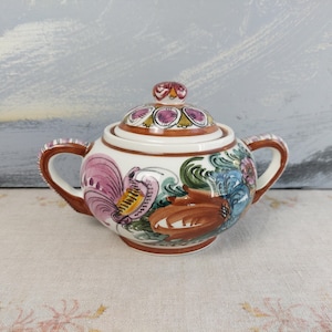 May include: A white ceramic sugar bowl with a lid, featuring hand-painted floral and rooster designs in pink, blue, and brown. The bowl has two brown handles and a matching knob on the lid. The sugar bowl is a decorative kitchenware item.