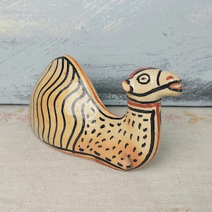 Puede incluir: Figura de camello de cerámica pintada a mano. El camello es beige con detalles negros y naranjas. La joroba tiene rayas verticales negras. La cabeza y el cuello tienen contornos negros y detalles rojos. Es un objeto decorativo.