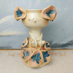 May include: A ceramic vase shaped like a stylized cat with closed eyes and blue and pink floral details.