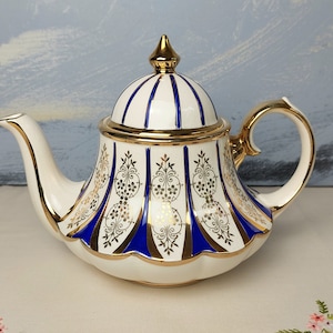 Op de afbeelding: Een witte theepot met een blauw en gouden ontwerp. De theepot heeft een koepelvormig deksel met een gouden knop en een breed, rond lichaam. Het ontwerp heeft blauwe strepen en gouden accenten.