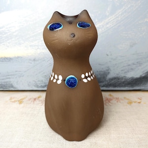 Puede incluir: Una figurita de cerámica marrón de un gato con ojos azules y un collar blanco y azul. El gato está mirando hacia arriba y tiene un diseño simple y caprichoso.