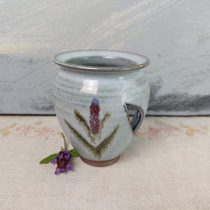 Könnte beinhalten: Ein hellblauer Keramikbecher mit bauchigem Körper und seitlichem Griffausschnitt. Die Außenseite zeigt ein gemaltes Blumenmotiv mit roter Mitte und grünen Blättern. Der Rand ist dunkelgrau und der Boden hat eine rotbraune Tonfarbe.