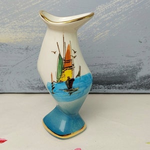 May include: A white ceramic vase with a blue and white seascape design. The vase has a gold trim around the top and bottom. The vase is shaped like a boat and has a small opening at the top.