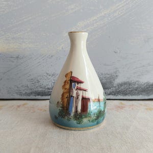 May include: A small, cream-colored ceramic vase with a tapered neck and a wide, rounded base. The exterior features a hand-painted scene of a coastal building with red roofs, blue accents, and green foliage, finished with a thin gold rim at the top.