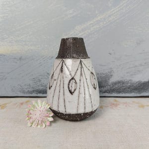 May include: A ceramic vase with a tapered neck and a rounded body. The neck and base are dark brown with a textured finish. The white body features a hand-painted black line pattern of vertical segments and leaf-like shapes encircling the center.
