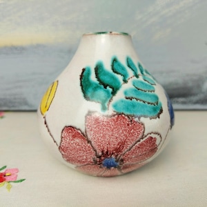 Puede incluir: Un pequeño jarrón de cerámica blanca con forma redondeada y un diseño floral. El jarrón está decorado con flores verdes, azules, rojas y amarillas.