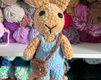 May include: A crocheted plush bunny toy wearing blue overalls and a brown satchel. The bunny has brown fur, black eyes, and a brown nose.