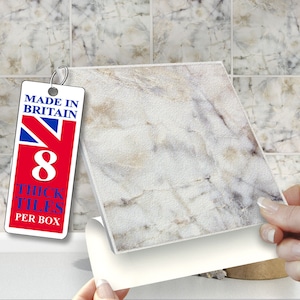 Op de afbeelding: Een close-up van een witte tegel met marmerlook en een subtiel beige patroon. De tegel wordt teruggetrokken om een kleverige zelfklevende achterkant te onthullen. De tegel is gelabeld met "Made in Britain" en "8 dikke tegels per doos".
