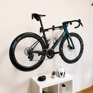Op de afbeelding: Een blauwgroene en zwarte racefiets gemonteerd aan de muur met een houten rek. De fiets heeft diepe carbon wielen, een racestuur en een zwart zadel. Onder de fiets staat een wit opbergmeubel met een witte helm, een plantje en een etui.