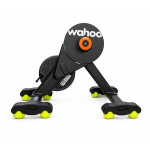 May include: A black Wahoo KICKR smart bike trainer featuring a circular housing with the white Wahoo logo and an orange center cap. The unit is supported by a sturdy frame with four black feet, each equipped with two bright yellow tennis balls.