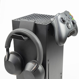 May include: A black Xbox Series X console with a black wireless controller and black over-ear headphones hanging on the side. The console has a black and white logo that says "Xbox Series X".
