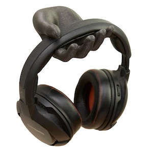 May include: A dark gray, textured, hand-shaped wall mount holding a pair of black over-ear headphones. The headphones feature black ear cushions with red interior accents and a black headband, resting securely on the fingers of the mount.