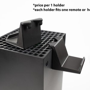 May include: Two black plastic holders for a video game console. The holders are designed to fit on the side of the console and can hold a remote or headphones. The text "*price per 1 holder *each holder fits one remote or headphones" is visible in the image.