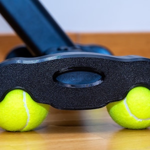 May include: A black plastic base component featuring the embossed text wahoo, designed with two circular cutouts that securely hold a pair of bright neon yellow tennis balls, providing a stable support structure for the connected black equipment arm.