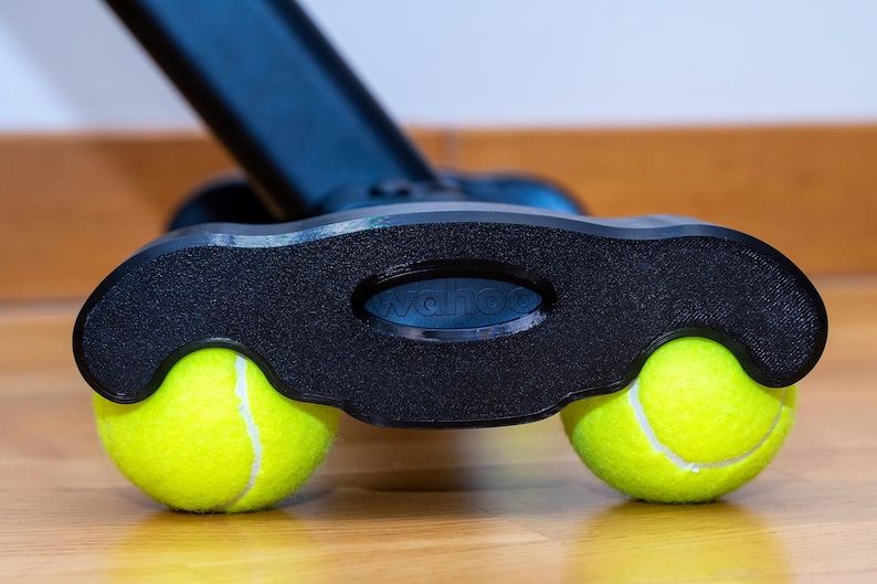 May include: A black plastic base component featuring the embossed text wahoo, designed with two circular cutouts that securely hold a pair of bright neon yellow tennis balls, providing a stable support structure for the connected black equipment arm.